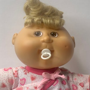Peut inclure: Une poupée Cabbage Patch Kid aux cheveux blonds portant une robe rose et blanche à cœurs. La poupée a une sucette dans la bouche.