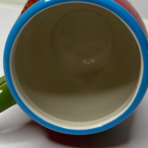 Puede incluir: Una taza de cer&aacute;mica con cuerpo rojo, borde azul y asa verde. El interior es beige. La taza tiene un dise&ntilde;o colorido en la parte superior. La taza est&aacute; dise&ntilde;ada para bebidas.