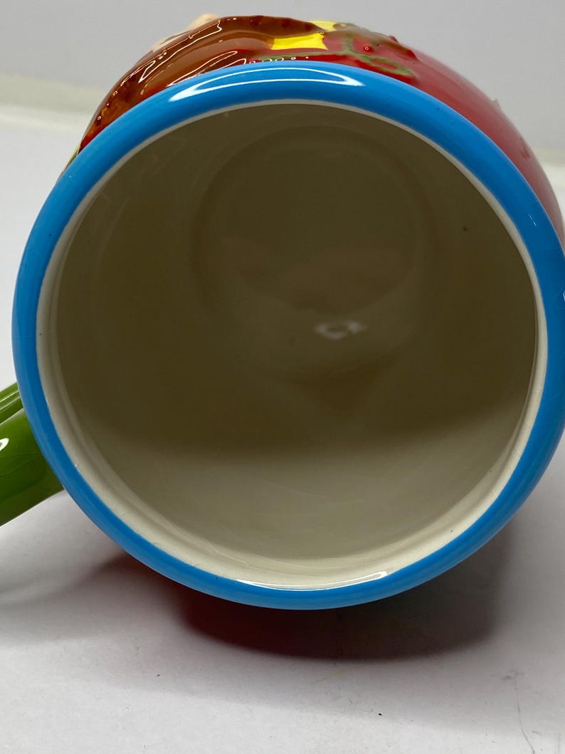 Puede incluir: Una taza de cer&aacute;mica con cuerpo rojo, borde azul y asa verde. El interior es beige. La taza tiene un dise&ntilde;o colorido en la parte superior. La taza est&aacute; dise&ntilde;ada para bebidas.