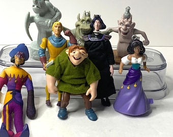 May include: A collection of colorful figurines featuring characters from a popular animated film. The figures include a gargoyle, a hunchback, and other characters in various poses. The figurines are made of plastic and have detailed features.