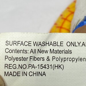 May include: A white tag with black text that reads "SURFACE WASHABLE ONLY.AI", "Contents: All New Materials", "Polyester Fibers & Polypropylen", "REG.NO.PA-15431(HK)", and "MADE IN CHINA". The tag is partially covering a yellow and red plaid pattern.