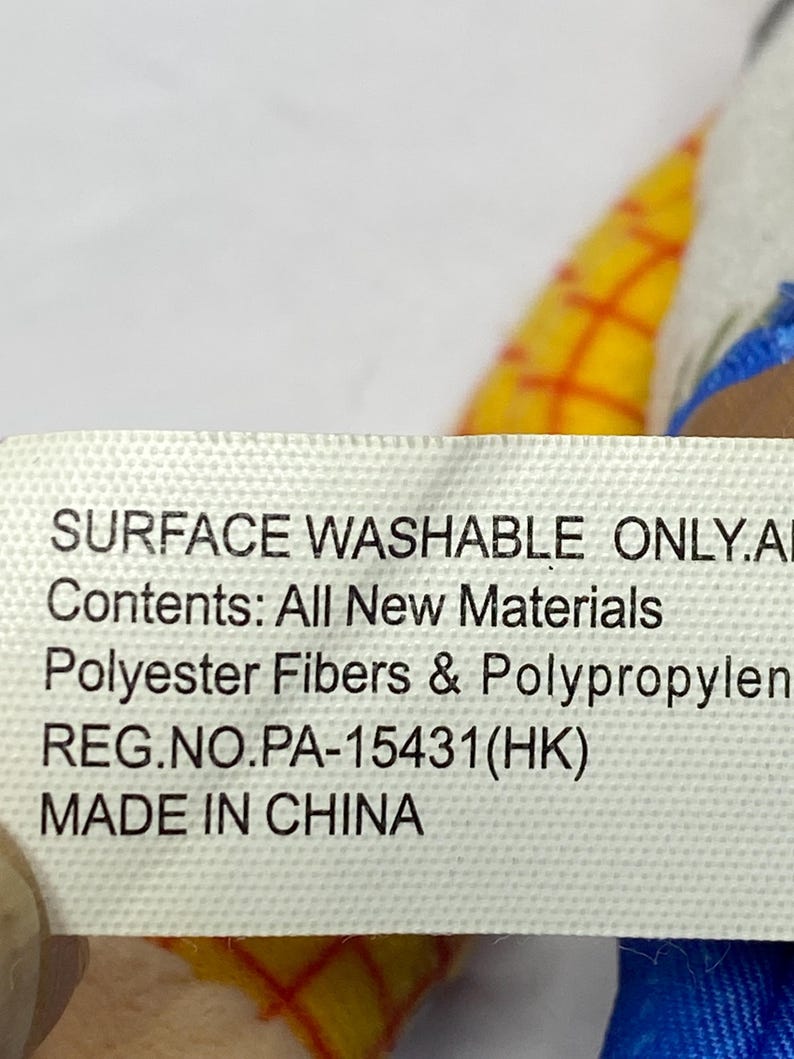 May include: A white tag with black text that reads "SURFACE WASHABLE ONLY.AI", "Contents: All New Materials", "Polyester Fibers & Polypropylen", "REG.NO.PA-15431(HK)", and "MADE IN CHINA". The tag is partially covering a yellow and red plaid pattern.