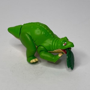 May include: A green dinosaur toy with a long neck and a leaf in its mouth. The toy has a textured body and is posed in a playful stance. The dinosaur's mouth is open, revealing a red interior.