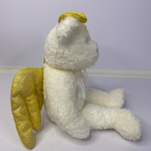 K&ouml;nnte beinhalten: Ein wei&szlig;er Pl&uuml;schteddyb&auml;r mit goldenen Glitzer-Engelsfl&uuml;geln und einem goldenen Heiligenschein. Der B&auml;r hat ein kleines, schillerndes Band um den Hals. Die Fl&uuml;gel sind auf dem R&uuml;cken des B&auml;ren positioniert. Der B&auml;r sitzt.