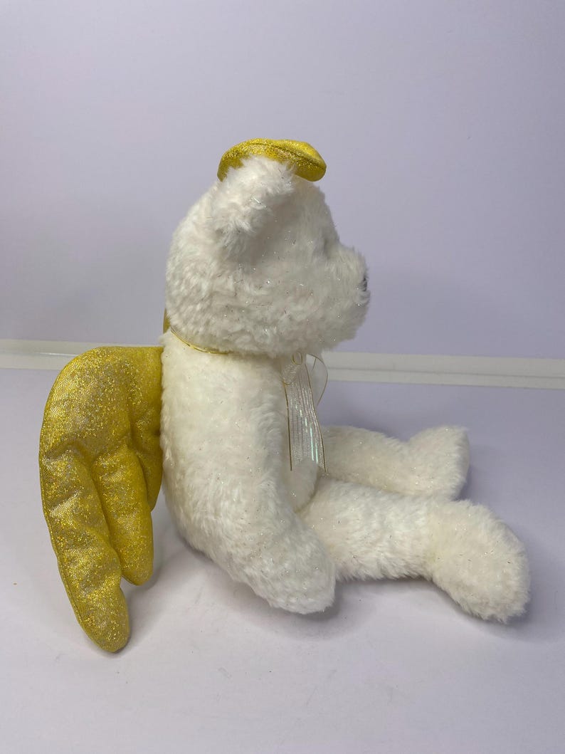 K&ouml;nnte beinhalten: Ein wei&szlig;er Pl&uuml;schteddyb&auml;r mit goldenen Glitzer-Engelsfl&uuml;geln und einem goldenen Heiligenschein. Der B&auml;r hat ein kleines, schillerndes Band um den Hals. Die Fl&uuml;gel sind auf dem R&uuml;cken des B&auml;ren positioniert. Der B&auml;r sitzt.