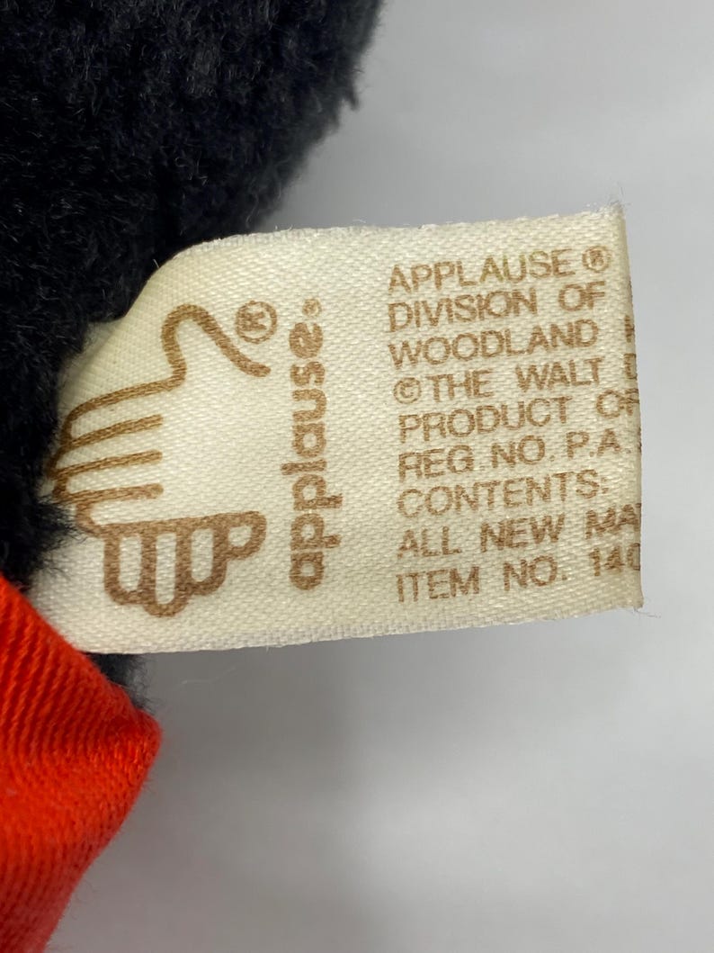 May include: A white tag with brown text that reads "APPLAUSE&reg; DIVISION OF WOODLAND &copy; THE WALT DISNEY PRODUCT OF REG. NO. P.A. CONTENTS: ALL NEW MATERIAL ITEM NO. 140". The tag is attached to a black plush toy.