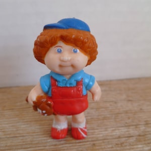 Op de afbeelding: Een plastic figuurtje van een jongen met rood haar, die een blauwe baseballpet, een blauw shirt, een rode overall en rode schoenen draagt. Hij houdt een bruine teddybeer vast.