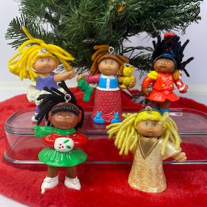 May include: Five vintage Cabbage Patch Kids Christmas tree ornaments. Each doll has yarn hair and is dressed in festive outfits, including a ballerina, a soldier, and a Christmas angel. The ornaments are displayed on a red tree skirt.