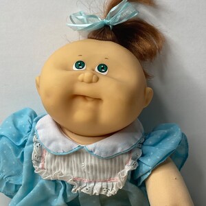 Puede incluir: Una muñeca Cabbage Patch Kid con cabello castaño atado con un lazo azul claro. La muñeca lleva un vestido azul claro con lunares blancos y un cuello blanco con ribete rosa.