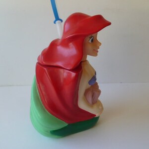 May include: A red and green plastic cup shaped like Ariel from The Little Mermaid. The cup has a straw and a sculpted image of Ariel's face and hair.