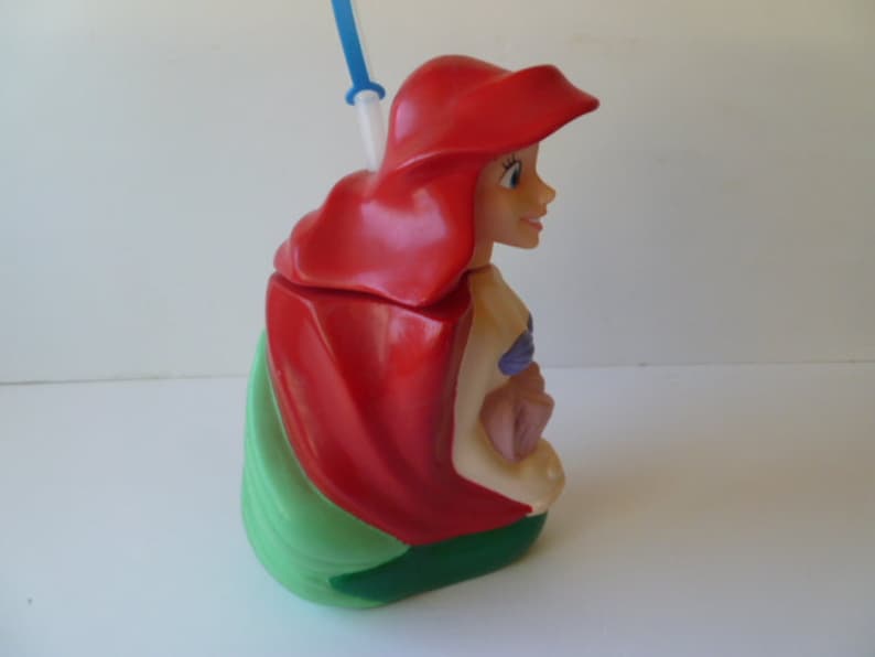 May include: A red and green plastic cup shaped like Ariel from The Little Mermaid. The cup has a straw and a sculpted image of Ariel's face and hair.