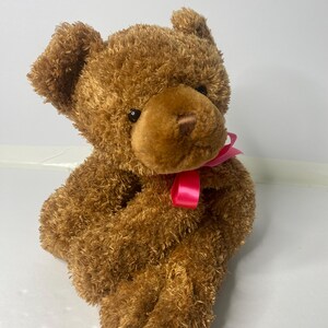 May include: A brown teddy bear with a pink bow sitting on a white surface.