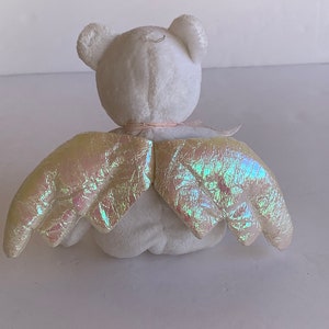 May include: White plush teddy bear with iridescent wings. The bear is sitting with its back to the camera. The wings are spread out behind the bear.