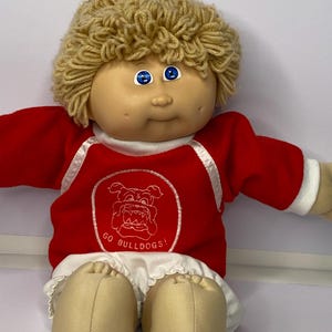 Könnte beinhalten: Eine Vintage Cabbage Patch Kid Puppe mit blondem Garnhaar und blauen Augen. Die Puppe trägt einen roten Sweatshirt mit weißem Besatz und dem Text "GO BULLDOGS!" auf der Vorderseite. Sie hat ein braunes Gesicht und Hände sowie weiße Rüschenshorts.