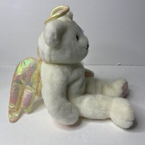 May include: White plush teddy bear with iridescent wings and a halo. The bear is sitting with its head turned to the side.