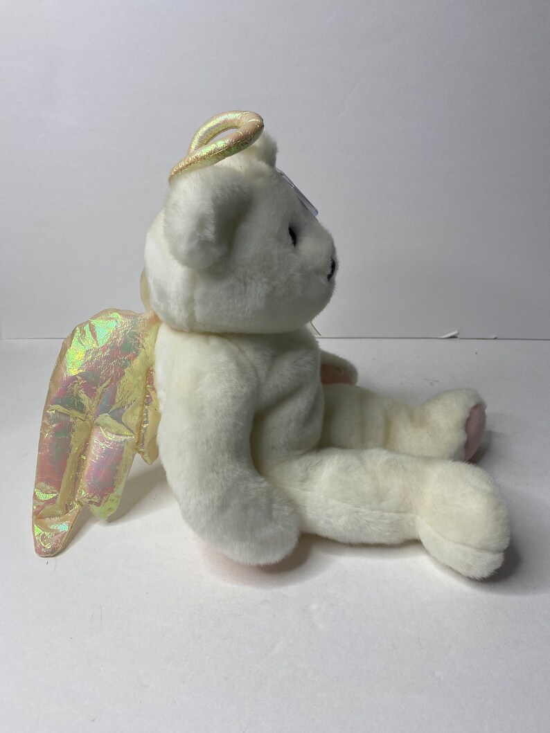 May include: White plush teddy bear with iridescent wings and a halo. The bear is sitting with its head turned to the side.