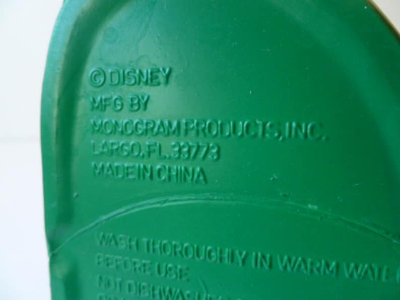 May include: Green plastic container with the text "DISNEY MFG BY MONOGRAM PRODUCTS, INC. LARGO, FL. 33773 MADE IN CHINA WASH THOROUGHLY IN WARM WATER BEFORE USE NOT DISHWASHER SAFE" printed on the bottom.