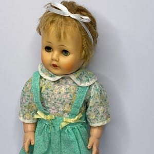 May include: A vintage doll with blonde hair and a white ribbon headband. The doll wears a floral print dress with a white collar and green suspender skirt. Yellow bows accent the skirt. The doll has painted facial features.