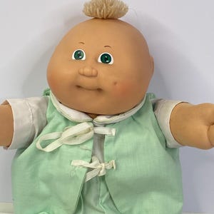 Op de afbeelding: Een vintage Cabbage Patch Kid pop met een kale kop, groene ogen en een kleine pluk lichtbruin haar. De pop draagt een lichtgroene outfit met witte randen en strikjes.