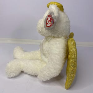 K&ouml;nnte beinhalten: Ein wei&szlig;er Pl&uuml;schteddyb&auml;r mit gelbem Heiligenschein und goldglitzernden Fl&uuml;geln. Der B&auml;r hat ein rot-wei&szlig;es Etikett an seinem Ohr. Der B&auml;r sitzt auf einer wei&szlig;en Oberfl&auml;che.