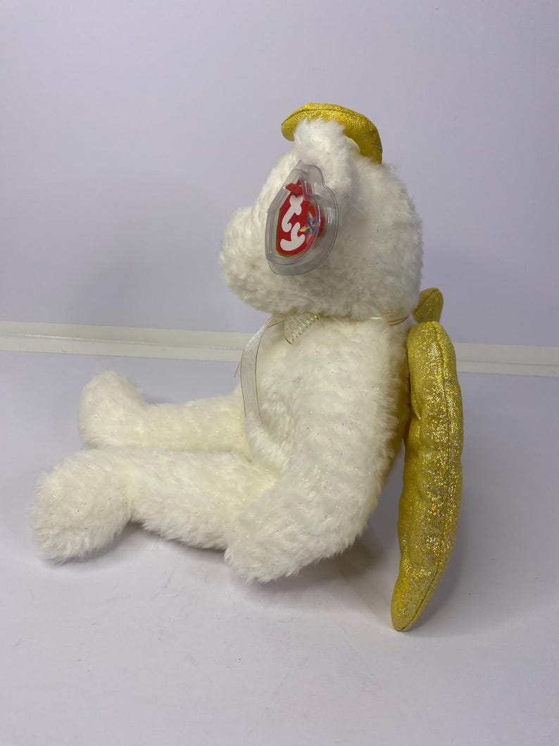 K&ouml;nnte beinhalten: Ein wei&szlig;er Pl&uuml;schteddyb&auml;r mit gelbem Heiligenschein und goldglitzernden Fl&uuml;geln. Der B&auml;r hat ein rot-wei&szlig;es Etikett an seinem Ohr. Der B&auml;r sitzt auf einer wei&szlig;en Oberfl&auml;che.