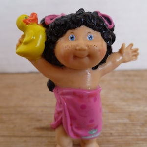 Può includere: Una bambola di plastica Cabbage Patch Kid con un pareo rosa e bianco a fantasia e che tiene un anatroccolo di gomma giallo.