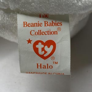 以下が含まれることがあります： 赤い文字で「The Beanie Babies Collection」と書かれた白いタグ。赤い星と赤いTyのハートのロゴが付いています。タグには「Halo」と登録商標記号と商標記号も書かれています。タグの下部には「Handmade in China」と書いてあります。