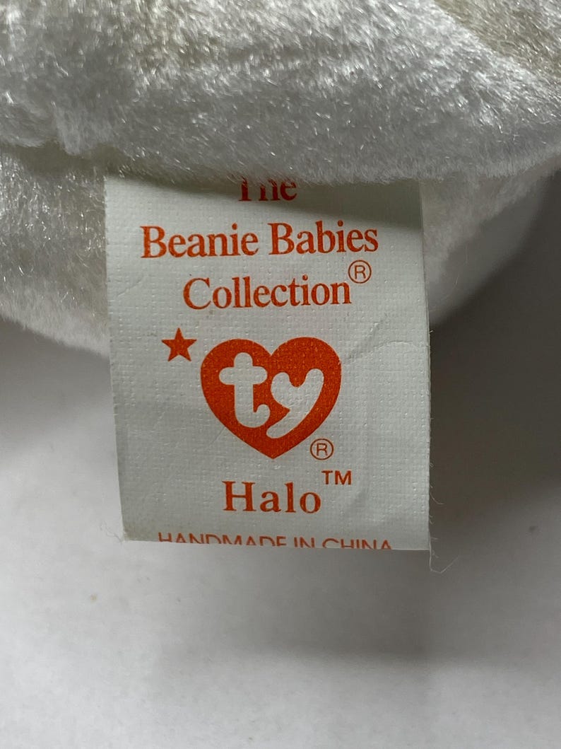 以下が含まれることがあります： 赤い文字で「The Beanie Babies Collection」と書かれた白いタグ。赤い星と赤いTyのハートのロゴが付いています。タグには「Halo」と登録商標記号と商標記号も書かれています。タグの下部には「Handmade in China」と書いてあります。