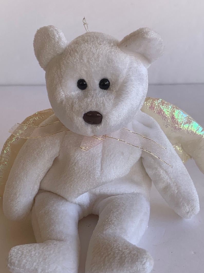 May include: White plush teddy bear with iridescent wings and a pink ribbon around its neck. The bear is sitting on a white surface.