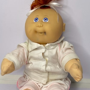 May include: A vintage Cabbage Patch Kid doll with fair skin, blue eyes, and red hair tied with a white ribbon. The doll is wearing a white and pink striped outfit with a collared shirt and matching trousers. The doll is sitting.