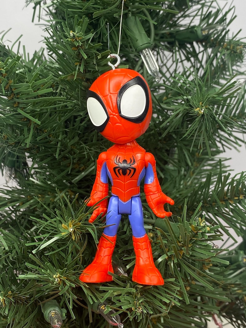 Puede incluir: Un adorno navide&ntilde;o de Spider-Man. El adorno es rojo, azul y negro, con grandes ojos blancos. La figura est&aacute; sujeta a un alambre plateado. El adorno est&aacute; colocado sobre un fondo de ramas verdes de &aacute;rbol de Navidad.