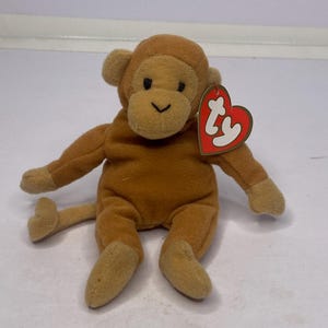 May include: A plush brown monkey toy with a tan face, ears, hands, and feet. The monkey has a small black stitched mouth and eyes. A red heart-shaped tag is attached to the monkey's ear.