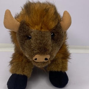 Peut inclure: Une peluche de bison avec une fourrure marron et beige, des cornes beiges et des sabots noirs. L'animal en peluche a un visage détaillé avec de petits yeux noirs et un museau brun foncé. Le bison est en position de repos.