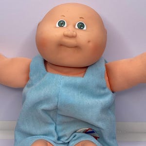 May include: A vintage Cabbage Patch Kid doll with a bald head, green eyes, and a light blue corduroy romper. The doll has a round face with rosy cheeks and a small, stitched mouth. The doll's arms and legs are made of a soft, peach-colored material.