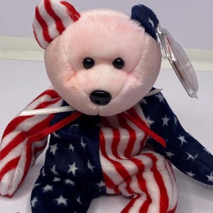 May include: A soft toy teddy bear with a patriotic design. The bear's body is split, with one side featuring red and white stripes and the other side displaying a blue background with white stars. The bear's head is pink, and it has black eyes and a black nose.