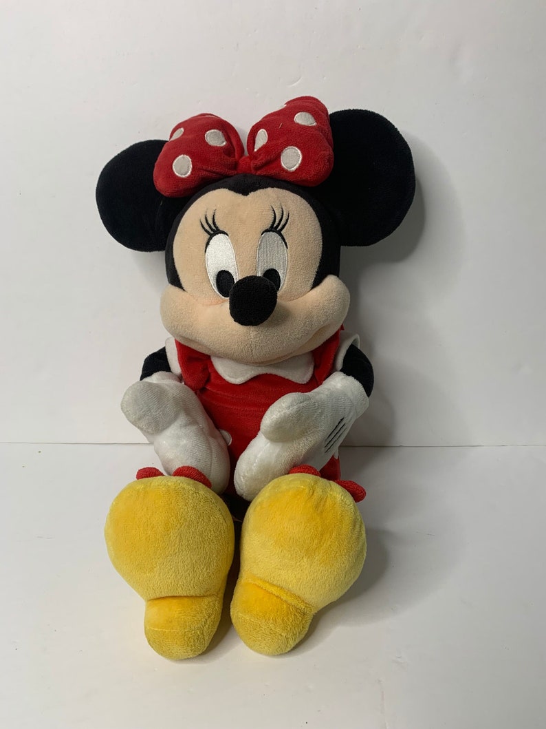 Pu&ograve; includere: Un giocattolo di peluche Minnie Mouse rosso e bianco con un fiocco rosso e scarpe gialle. Il giocattolo &egrave; seduto con le gambe incrociate.