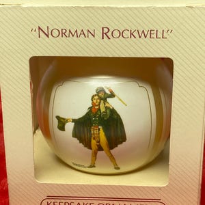 May include: A Hallmark Keepsake Ornament with a Norman Rockwell illustration. The white sphere ornament depicts a man and child. The beige box has the text "Norman Rockwell" and "Keepsake Ornament".