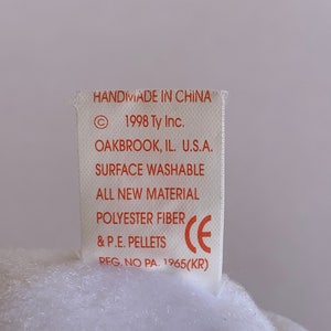 May include: A white tag with red text that reads "Handmade in China", "1998 Ty Inc.", "Oakbrook, IL. U.S.A.", "Surface Washable", "All New Material", "Polyester Fiber", "& P.E. Pellets", "Reg. No PA. 1965(KR)" and a CE mark.
