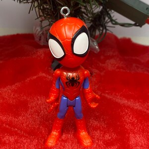 Puede incluir: Figura de Spider-Man con un traje rojo y azul, un emblema de ara&ntilde;a negro y ojos blancos. La figura tiene un lazo plateado para colgar. Sobre un fondo rojo y un &aacute;rbol de Navidad verde.