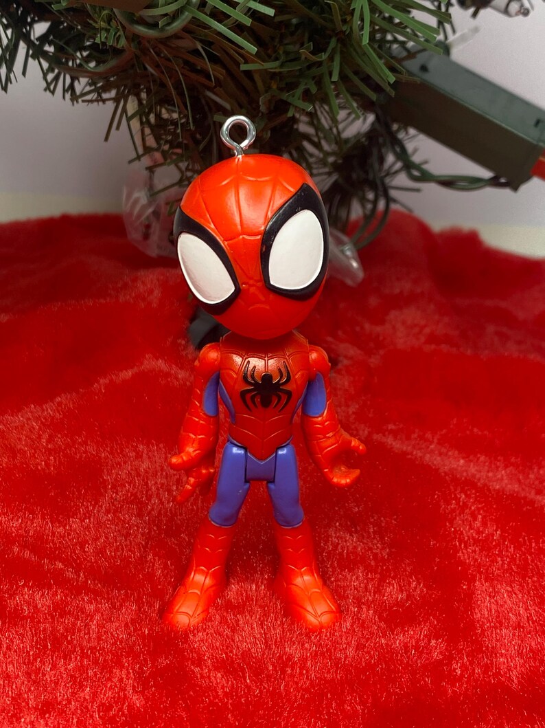 Puede incluir: Figura de Spider-Man con un traje rojo y azul, un emblema de ara&ntilde;a negro y ojos blancos. La figura tiene un lazo plateado para colgar. Sobre un fondo rojo y un &aacute;rbol de Navidad verde.