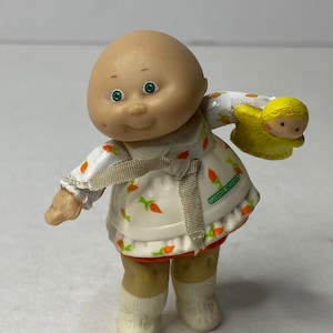 Op de afbeelding: Een vintage Cabbage Patch Kids beeldje. De pop heeft een kale kop, groene ogen en draagt een witte schort met oranje wortelontwerpen. Het houdt een gele pop vast. Het beeldje draagt witte sokken en schoenen.
