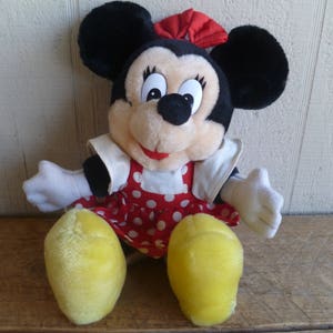 Op de afbeelding: Een pluche speelgoed van Minnie Mouse met een rood-wit geblokt jurkje met een rode strik. Het speelgoed heeft zwarte oren, gele schoenen en een glimlachend gezicht.
