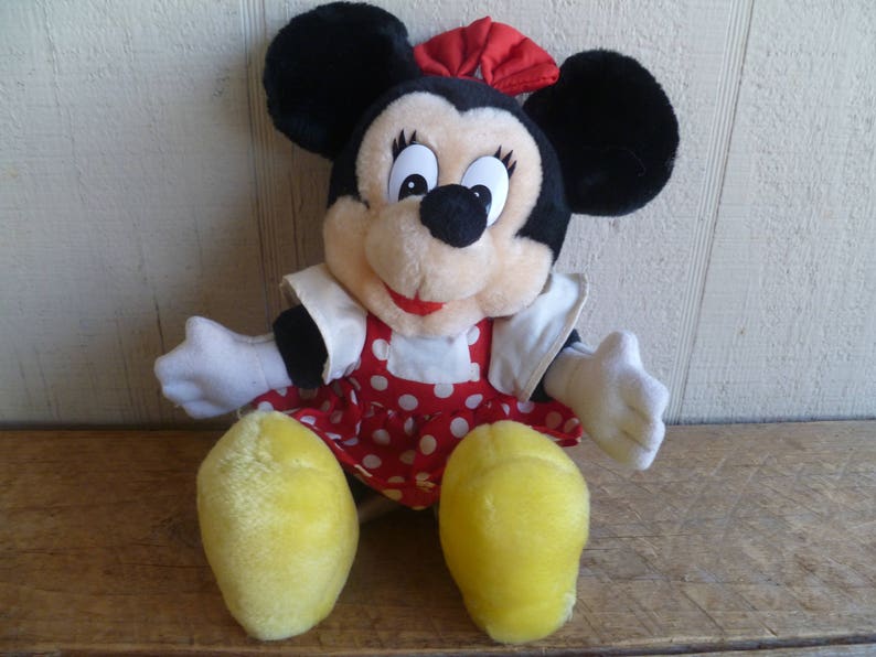 Op de afbeelding: Een pluche speelgoed van Minnie Mouse met een rood-wit geblokt jurkje met een rode strik. Het speelgoed heeft zwarte oren, gele schoenen en een glimlachend gezicht.