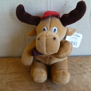 May include: A brown plush moose wearing a red and black plaid hat. The moose is holding a small plaid scarf in its paws. The moose is sitting on a wooden surface. The tag on the moose says "Things".