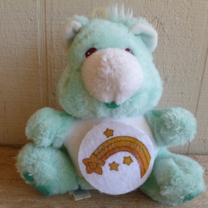 Może przedstawiać: Jasno niebieski pluszowy Miś Kubuś Puchatek z białym łatką na brzuchu, na której znajduje się tęcza i gwiazdki. Miś ma biały pyszczek i czerwone oczy.