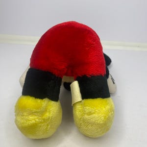 May include: A soft toy depicting Mickey Mouse's red shorts, black legs, and yellow feet. The toy is made of a soft, textured fabric and is positioned upside down. A small tag is attached to one leg.