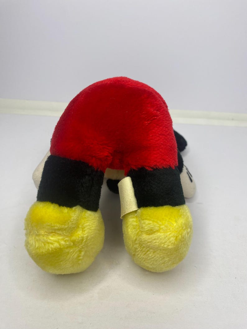May include: A soft toy depicting Mickey Mouse's red shorts, black legs, and yellow feet. The toy is made of a soft, textured fabric and is positioned upside down. A small tag is attached to one leg.
