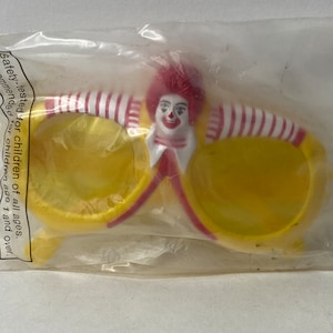 May include: Yellow plastic sunglasses with red and white stripes. The sunglasses have a cartoon image of Ronald McDonald on the front.