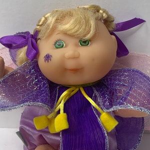 Puede incluir: Una muñeca Cabbage Patch Kid vestida con un traje de hada morado y rosa con detalles amarillos. La muñeca tiene el pelo rubio, los ojos verdes y una pegatina de flor en la mejilla.
