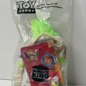 May include: A green Buzz Lightyear plush toy from the Disney Pixar movie Toy Story. The toy is wearing a Burger King uniform with the Burger King logo and the words "See the movie at theaters everywhere." The toy is packaged in a clear plastic bag with a Burger King logo.